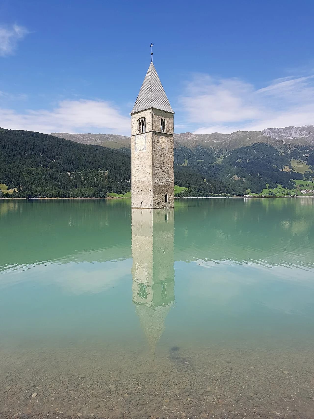 Due facciate del campanile di Curon, con muratura, quadranti e riflesso nell’acqua
